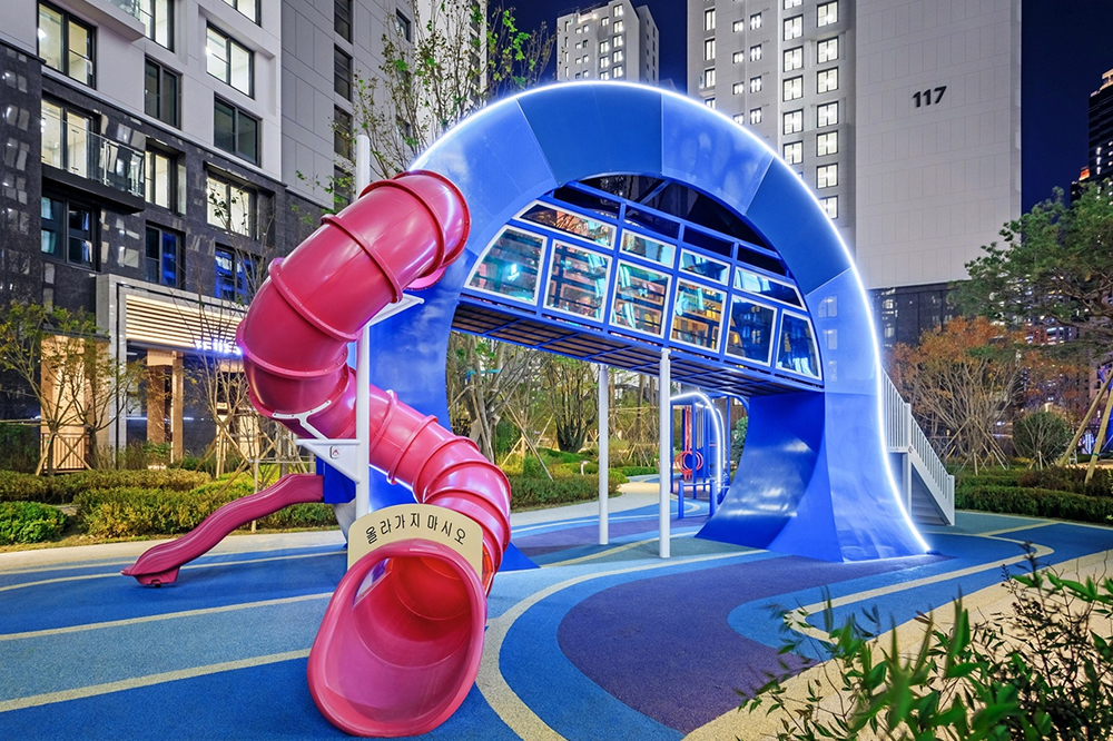 Modern playground at Jamsil Raemian I’Park with a large curved blue arch structure and red slide, illuminated at night and surrounded by apartment buildings.