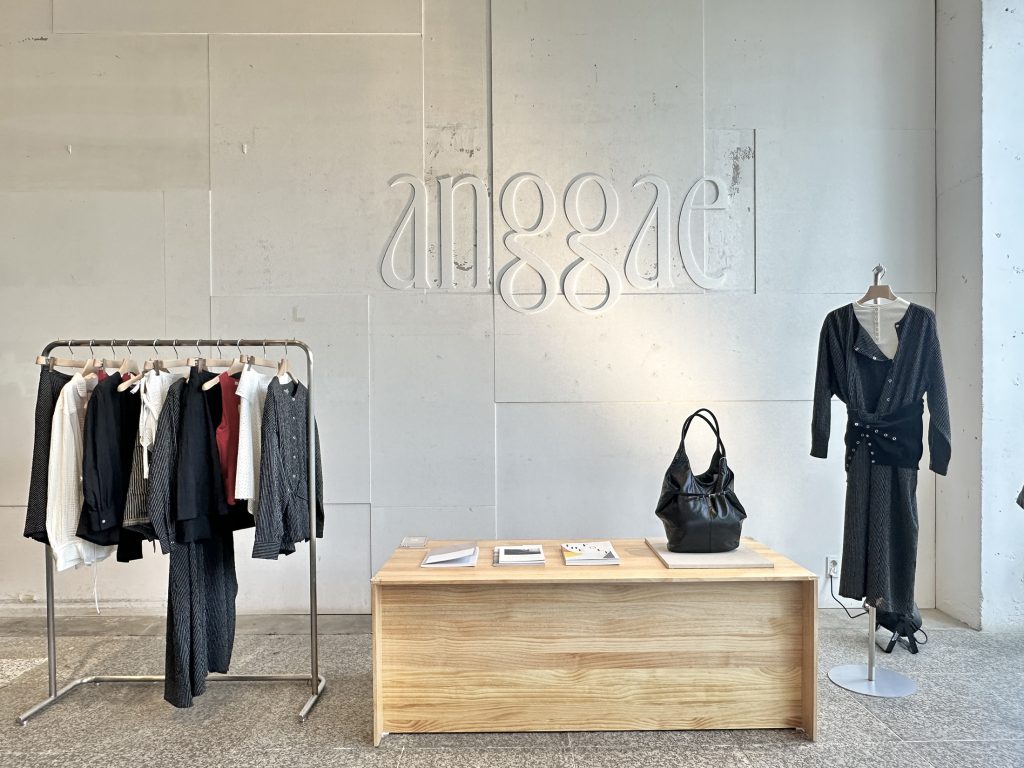 Interior view of anggae’s 2026 Spring pop-up store at Beaker Cheongdam, featuring neutral-toned womenswear displayed on a clothing rack, a wooden table with accessories, and the brand logo on a minimalist wall.