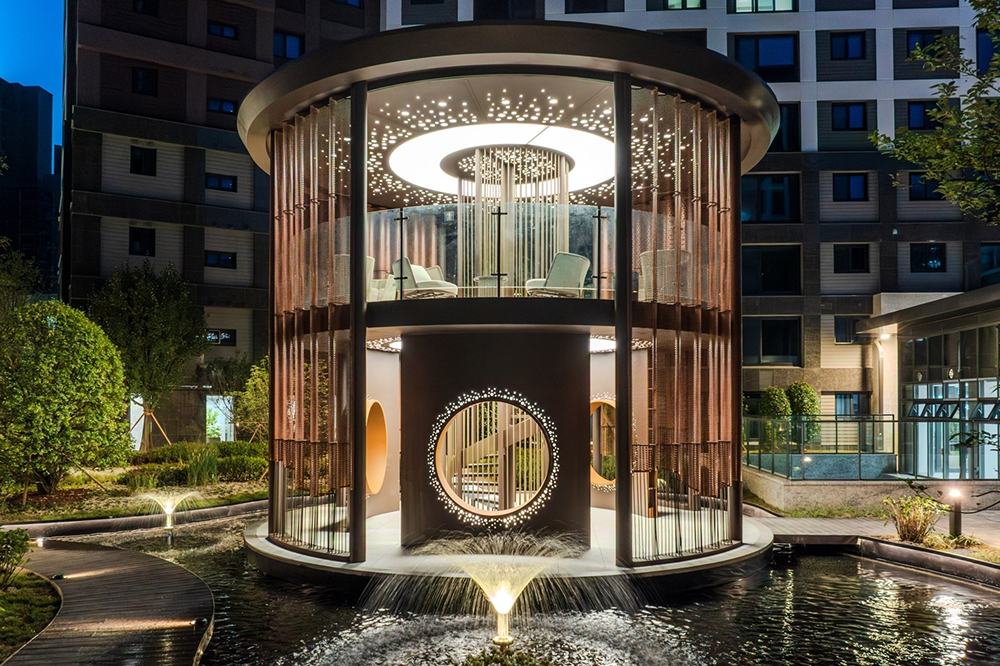 Circular two-story pavilion at Raemian One Perla featuring illuminated ceiling details, seating areas, and surrounding water features at night.