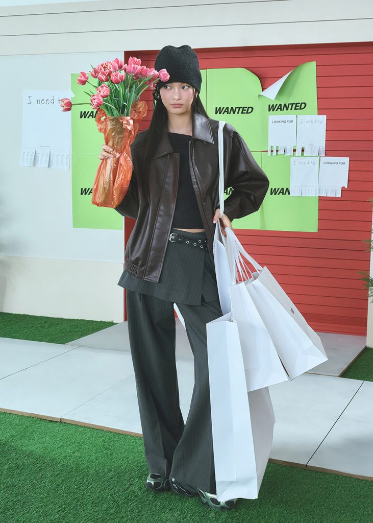 Model wearing a brown leather jacket layered over a black cropped top with a skirt-over-pants combination from KUHO PLUS SS 2026 collection.