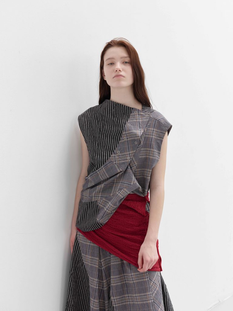 Model wearing a striped sleeveless top layered with a plaid overlay and a red wrap detail styled at the waist from anggae’s SS 2026 collection.