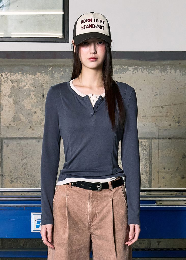 Model wearing a grey Henley-style long-sleeve top layered over a white inner shirt with beige trousers and a baseball cap from 8SECONDS SS 2026 collection.