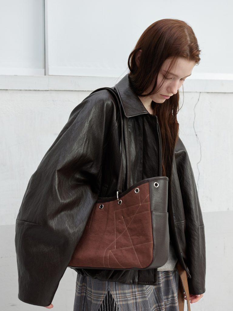 Model wearing a brown faux leather blouson jacket from anggae’s 2026 Spring collection, styled with a brown Gardener Bag featuring contrast stitching and long shoulder straps.