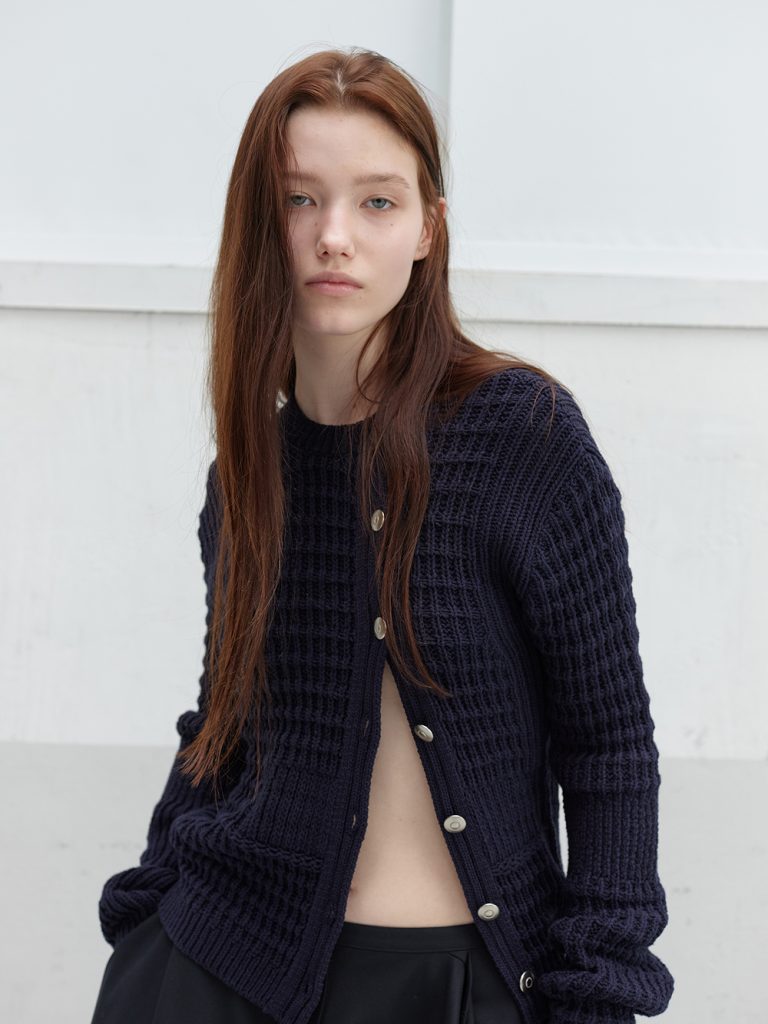 Model wearing a navy waffle-knit cardigan from anggae’s 2026 Spring collection, styled with relaxed black pants in a minimalist setting.