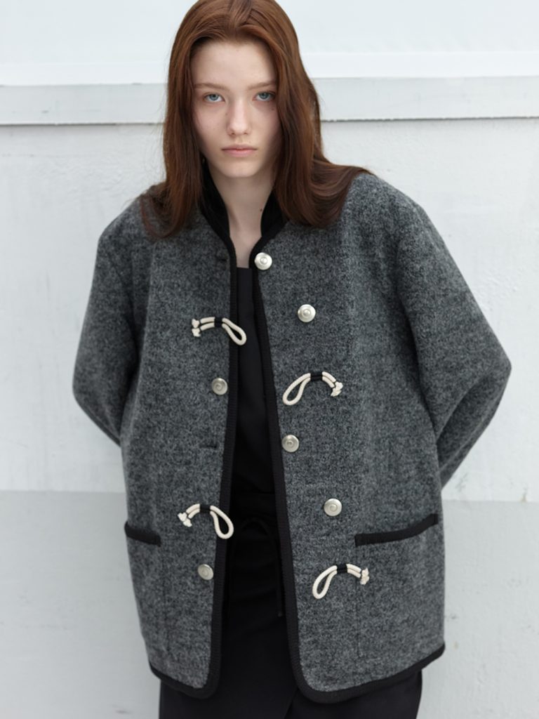 Model wearing a grey tweed Gardener Jacket from anggae’s 2026 Spring collection, detailed with rope-button fastenings, black trim, and front patch pockets.