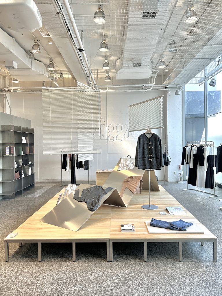 Wide interior view of anggae’s 2026 Spring pop-up store at Beaker Cheongdam, featuring a central wooden display table with garments and accessories, minimalist metal fixtures, and hanging clothing racks in a bright, open space.