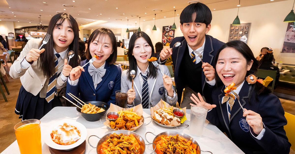 A group of visitors wearing school uniforms sit around a table enjoying tteokbokki and various snack dishes during Everland’s School of Spice event