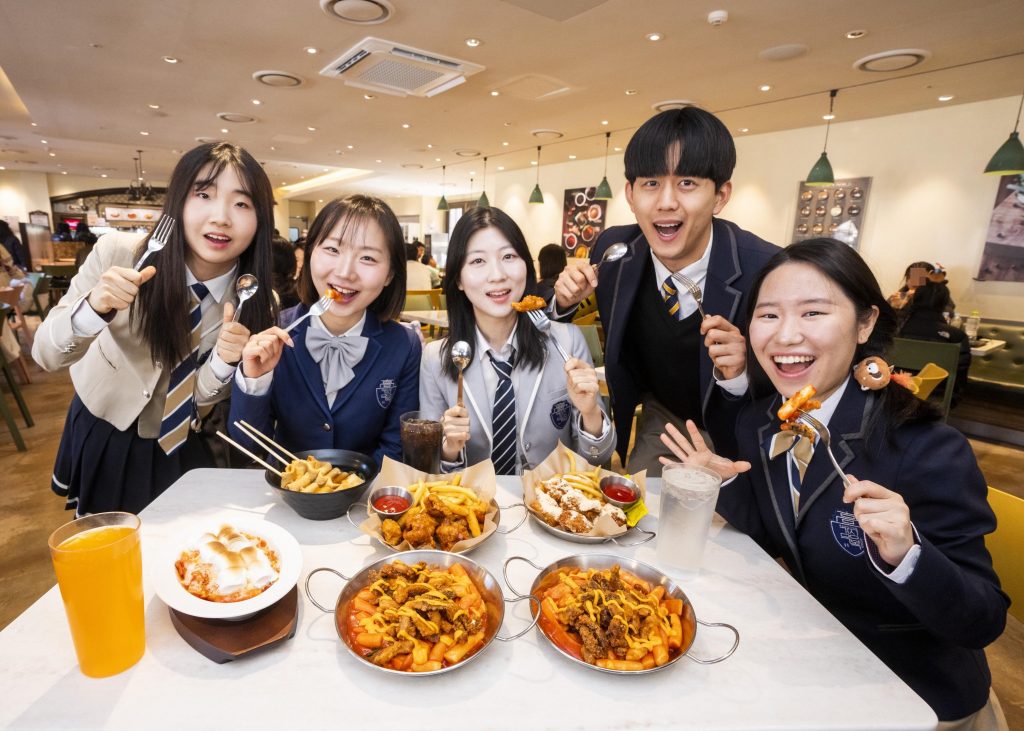 A group of visitors wearing school uniforms sit around a table enjoying tteokbokki and various snack dishes during Everland’s School of Spice event