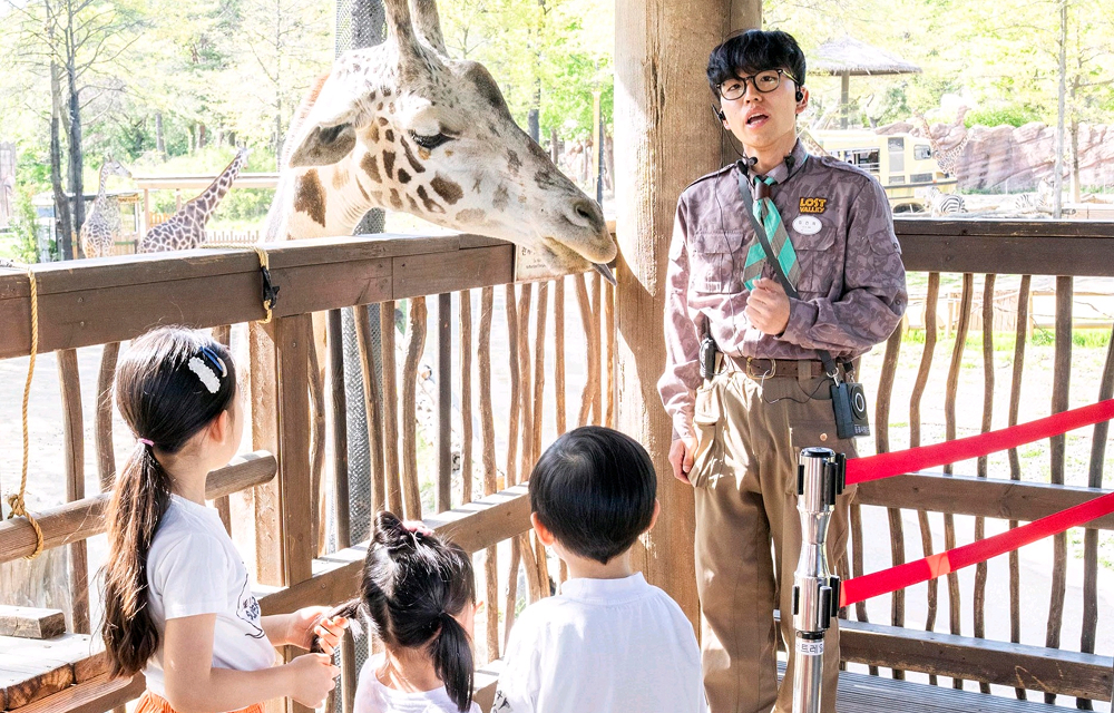 Children stand beside a Lost Valley guide at Everland while watching a giraffe lean over a wooden fence during an Ever Kids Club animal adventure program.