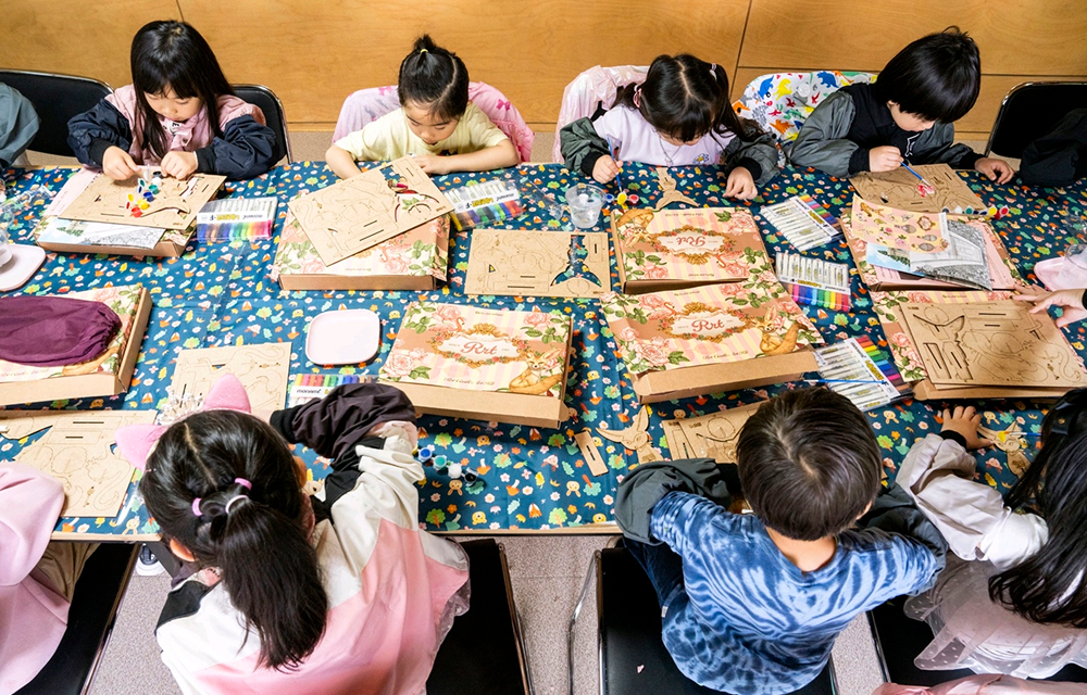 Children sit around a large table during an Ever Kids Club workshop at Everland, painting and decorating wooden craft boards with colorful markers and materials.
