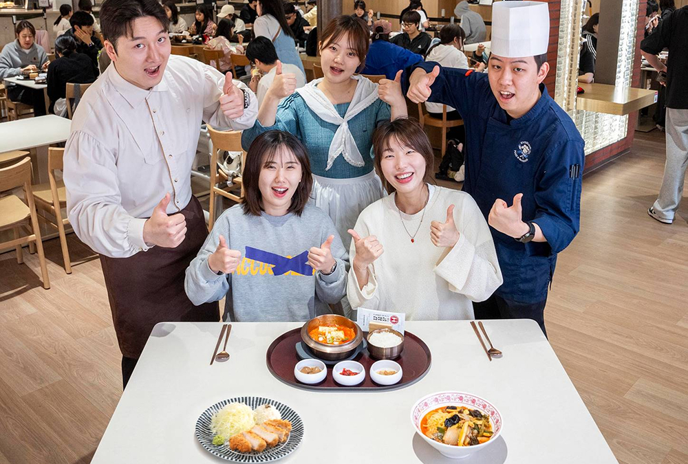 Visitors and Everland restaurant staff pose with three signature menus on a table, including kimchi stew, spicy seafood noodle soup, and handmade roast pork cutlet. 