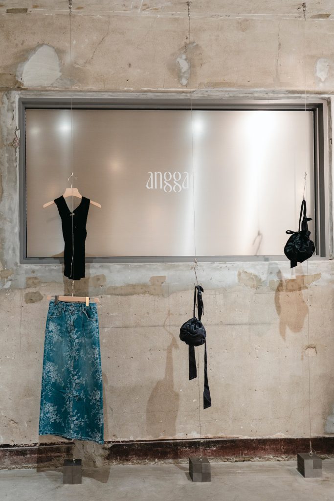 Clothing and accessories from the ‘anggae’ 26SS collection displayed on suspended hangers against a textured wall.