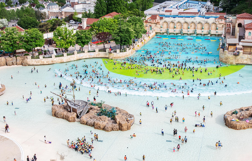 Wide view of visitors playing in Caribbean Bay’s outdoor Wave Pool with the water park’s Mediterranean-style buildings in the background.