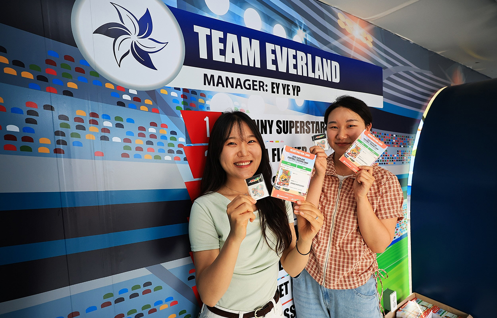 Two visitors smile while holding mission sheets and small rewards in front of a Team Everland football-themed wall.