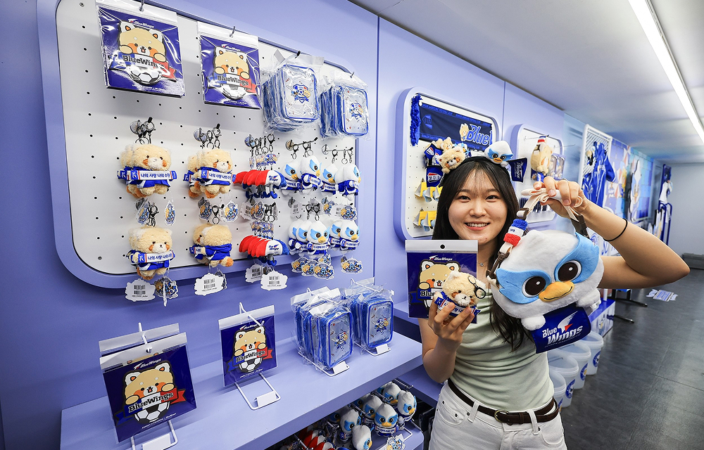 A visitor holds character merchandise inside a blue-themed goods shop displaying Suwon Samsung Bluewings and Everland collaboration items.