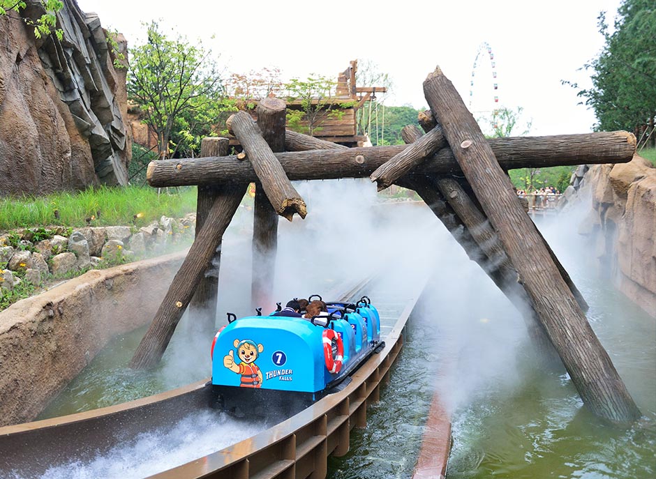A blue Thunder Falls ride vehicle moves through a water-filled track under large wooden structures, surrounded by mist and splashing water.