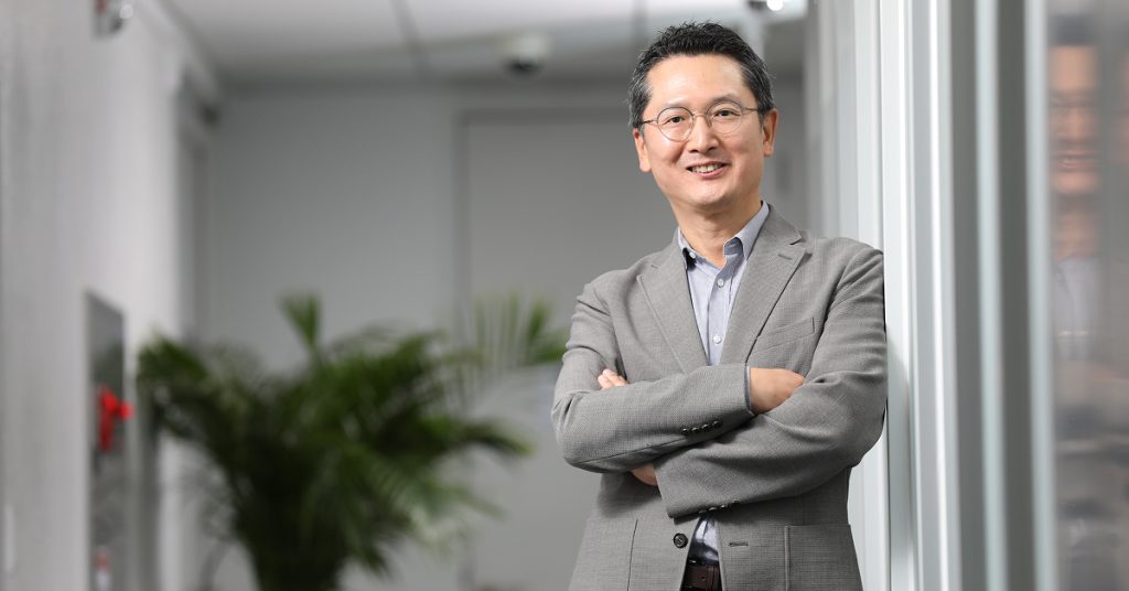 Jusung Park stands indoors with his arms crossed, smiling near a glass wall.