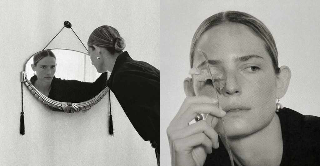 Model wearing a dark look from The Aperture’s Fall 2026 collection leaning toward a vintage wall mirror in a black-and-white photograph.