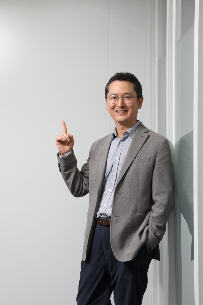Jusung Park stands indoors, smiling and pointing upward beside a glass wall.