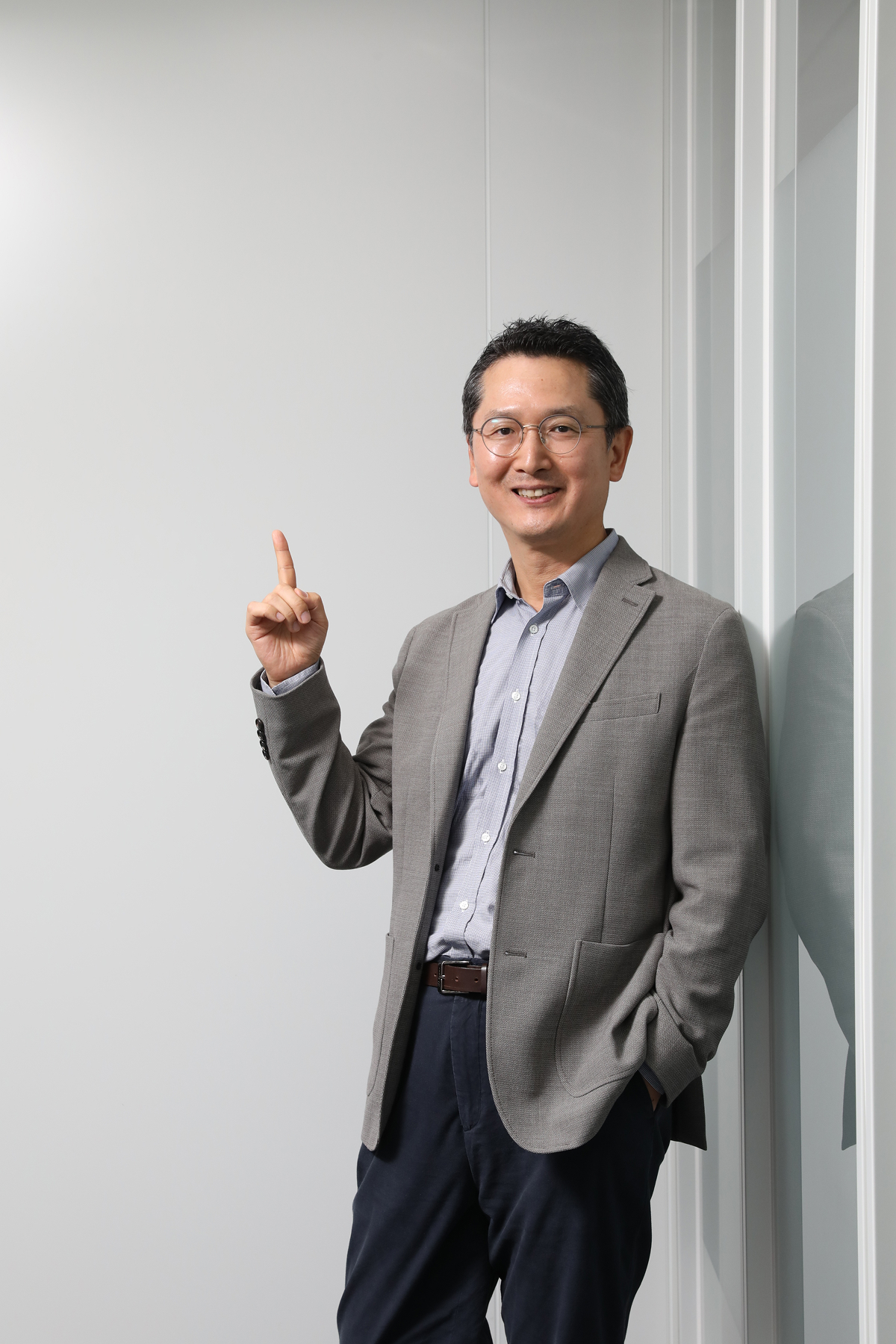 Jusung Park stands indoors, smiling and pointing upward beside a glass wall.