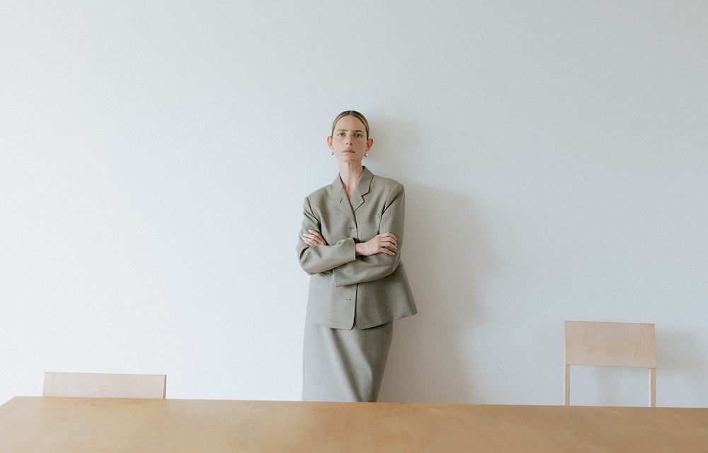 Model wearing a tailored gray jacket and matching H-line skirt standing against a white wall behind a long wooden table.
