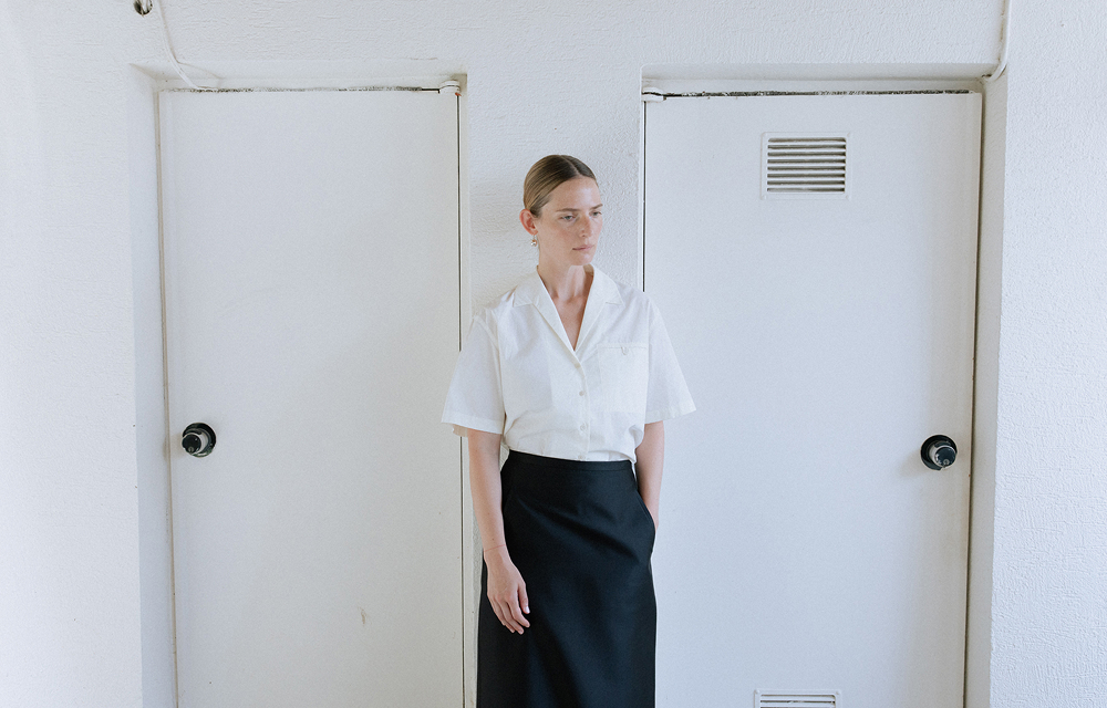 Model wearing a white short-sleeve shirt and long black H-line skirt standing between two white doors.