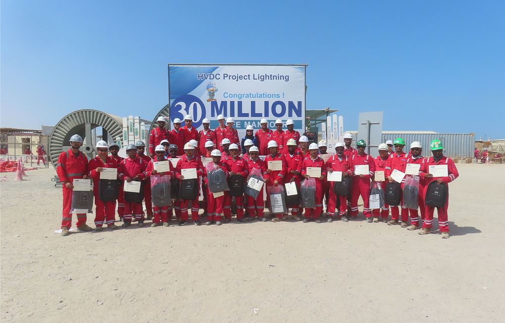 UAE HVDC project employees in red protective clothing pose with certificates and gifts in front of a banner celebrating 30 million work hours without a lost-time injury.