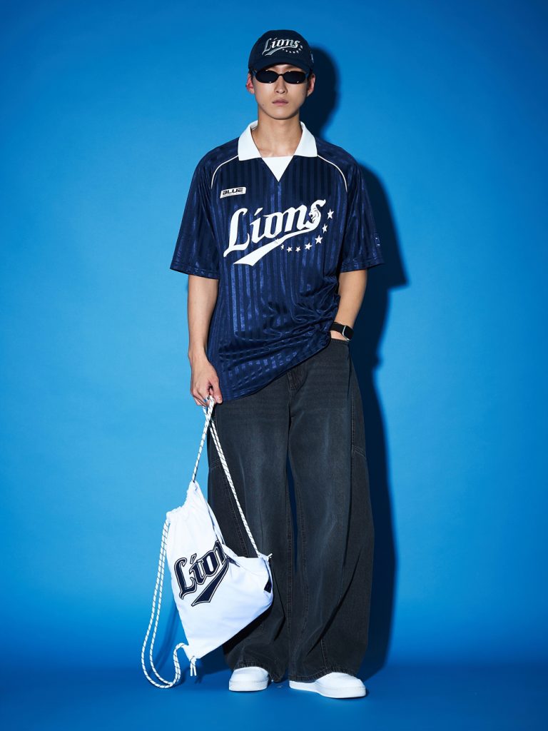 Model wearing a navy Samsung Lions jersey and cap with black wide-leg jeans, white sneakers, sunglasses, and a white Lions drawstring bag against a blue backdrop.