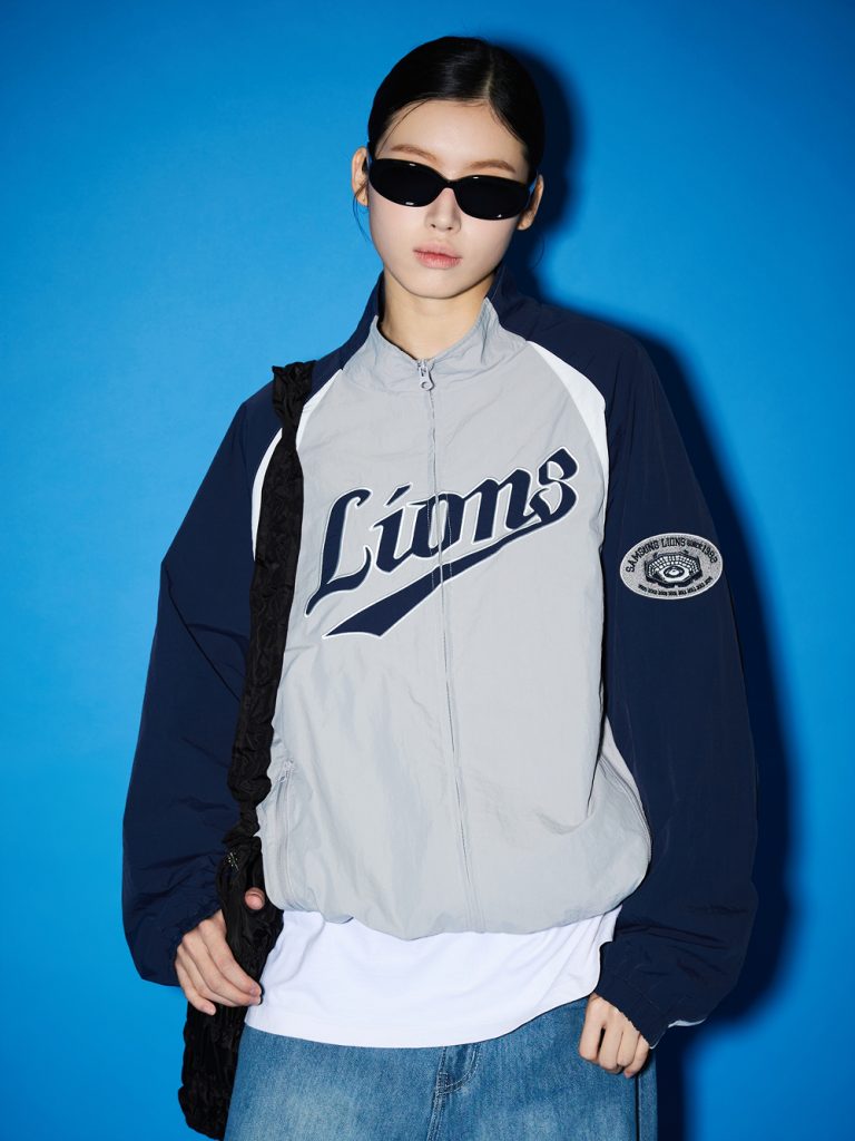 Model wearing a gray-and-navy Samsung Lions color-block windbreaker over a white T-shirt and jeans, styled with black sunglasses and a shoulder bag against a blue backdrop.