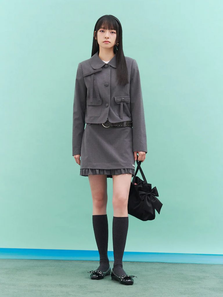 Model wearing a gray cropped round-neck jacket with a matching pleated mini skirt, black knee-high socks and black shoes, holding a black ribbon bag against a mint-green backdrop.