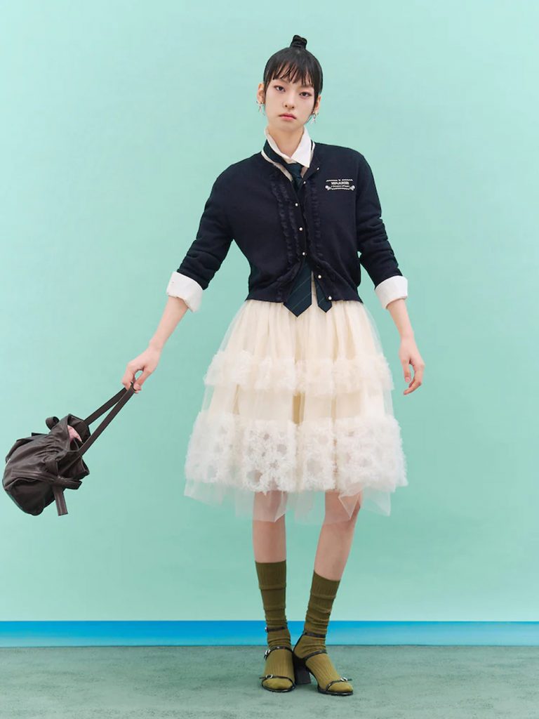 Model wearing a navy ribbon-detail cardigan over a white collared top and layered white tulle skirt, holding a dark bag against a mint-green backdrop.