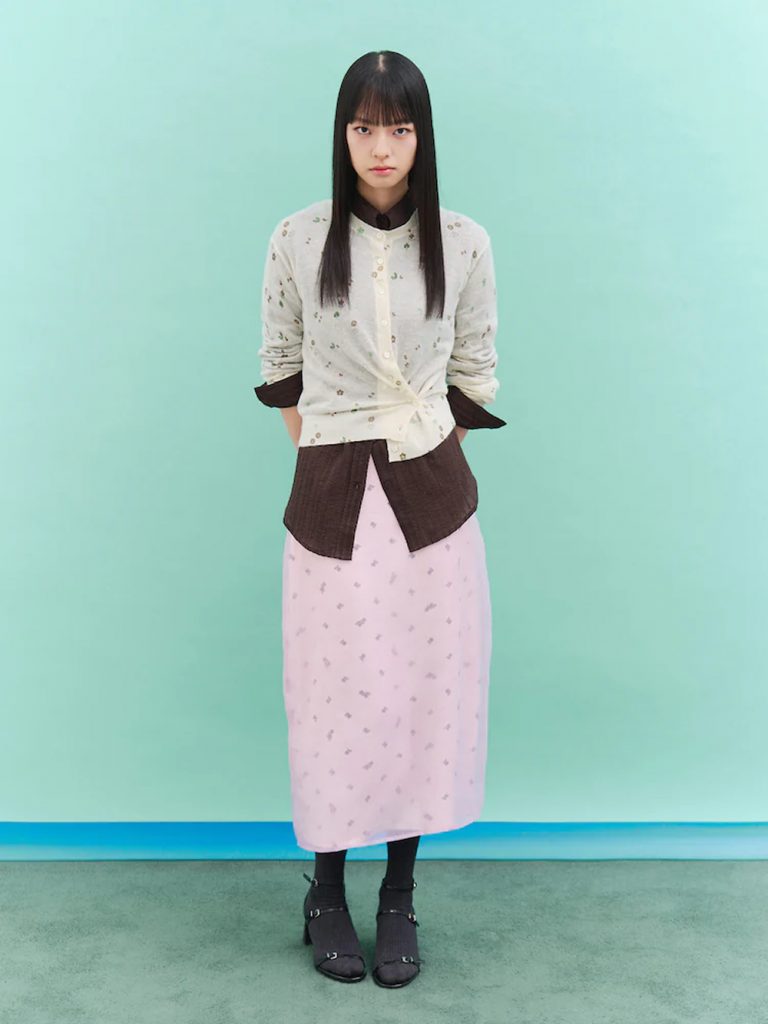 Model wearing an ivory heart-clover print cardigan layered over a brown top and pale pink skirt, styled with dark socks and shoes against a mint-green backdrop.