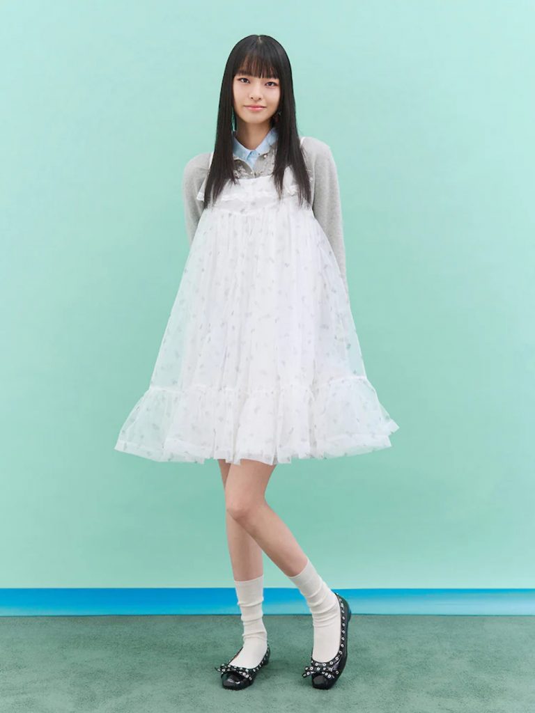 Model wearing a white floral-pattern layered tulle dress over a light blue collared top, paired with white socks and black shoes against a mint-green backdrop.