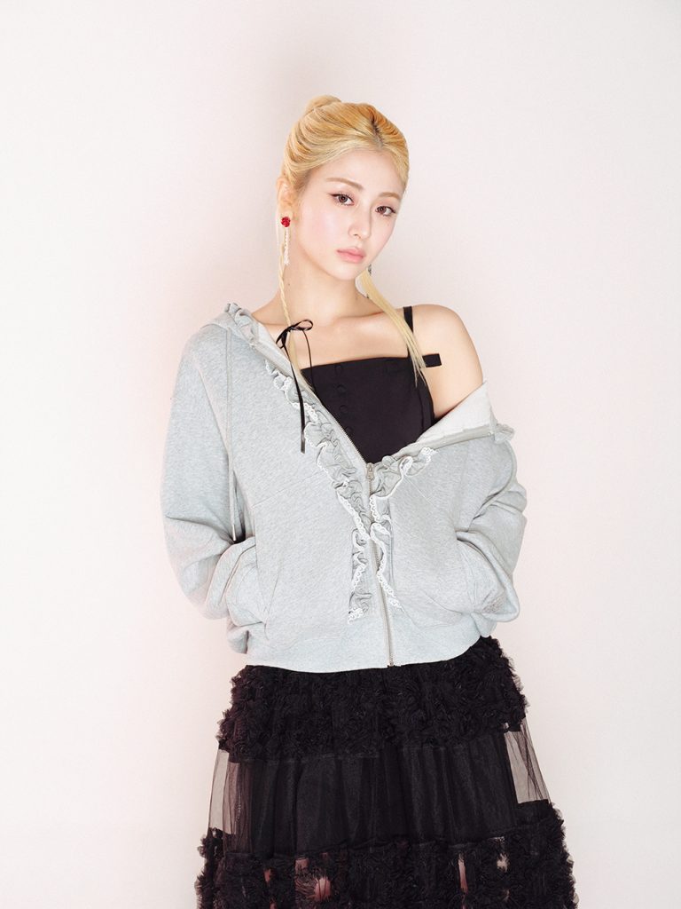 A model with blonde hair wears a gray zip-up hoodie with ruffle trim over a black top and a layered black tulle skirt, finished with long statement earrings against a pale backdrop.