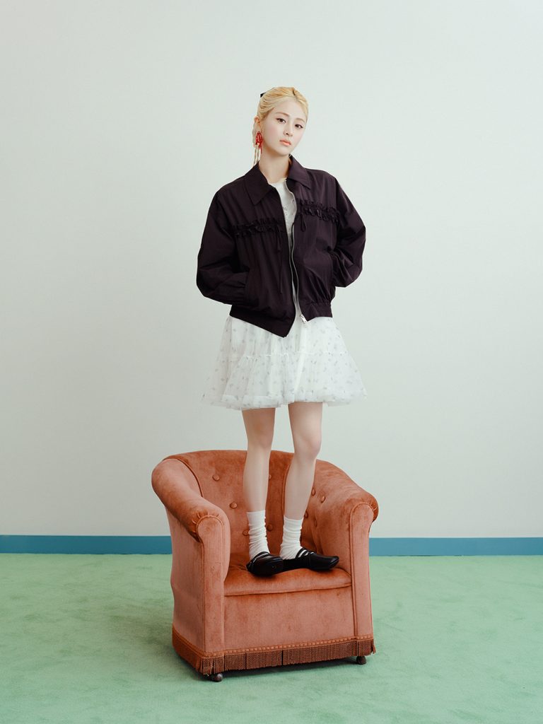 A model stands on an orange armchair wearing a dark ruffle-detail zip-up jumper over a short white tulle dress, paired with white socks and black shoes in a mint-green studio setting.