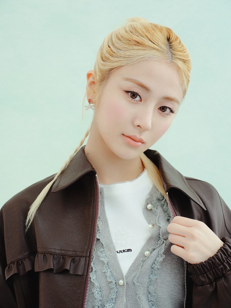 Close-up portrait of a model wearing a dark brown faux-leather blouson over a gray ruffle-trim cardigan with pearl-style buttons and a white top, against a pale mint-green background.