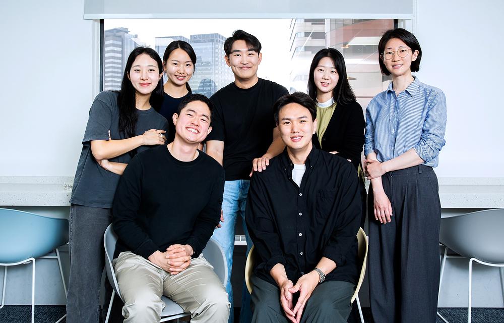 Seven members of Samsung C&T’s Development 1 Group East and Wind Teams pose together indoors in front of a large window.