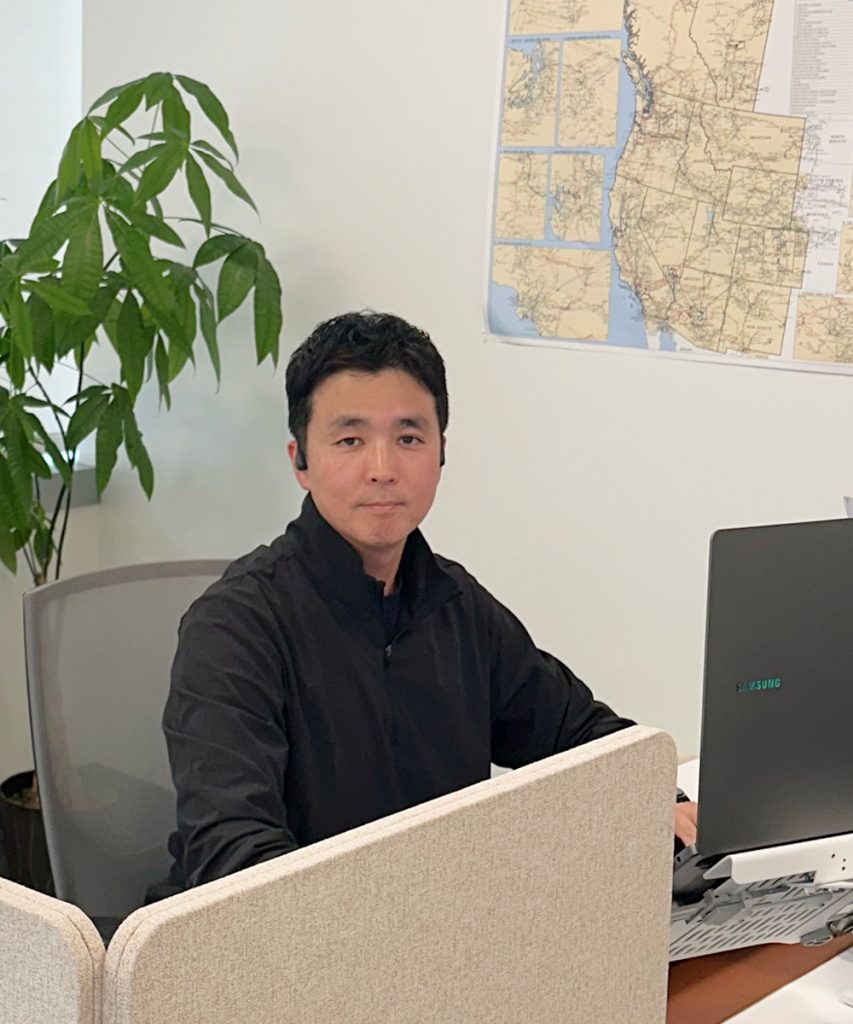 HyungHoon Lim sits at a desk in an office, looking at the camera with plants, windows, and a wall map behind him.