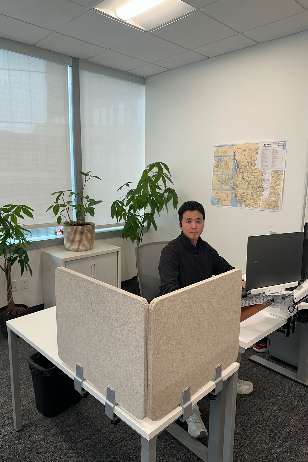 HyungHoon Lim sits at a desk in an office, looking at the camera with plants, windows, and a wall map behind him.