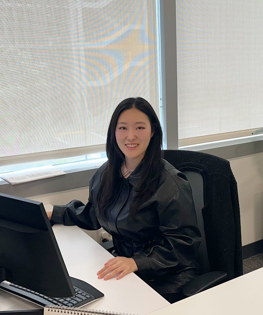 Jie Eun Chung sits at a desk in an office, smiling at the camera with windows, plants, and workstations around her.
