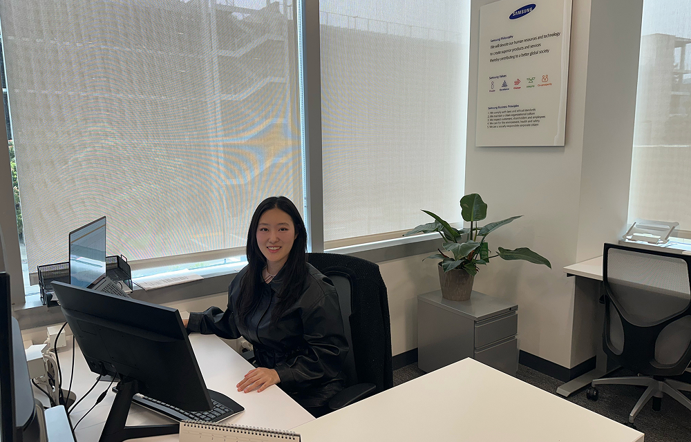 Jie Eun Chung sits at a desk in an office, smiling at the camera with windows, plants, and workstations around her.
