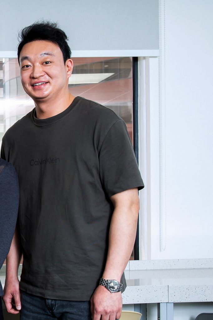 Tai Seok Kim stands indoors in front of a large window, smiling at the camera in a dark T-shirt and jeans.