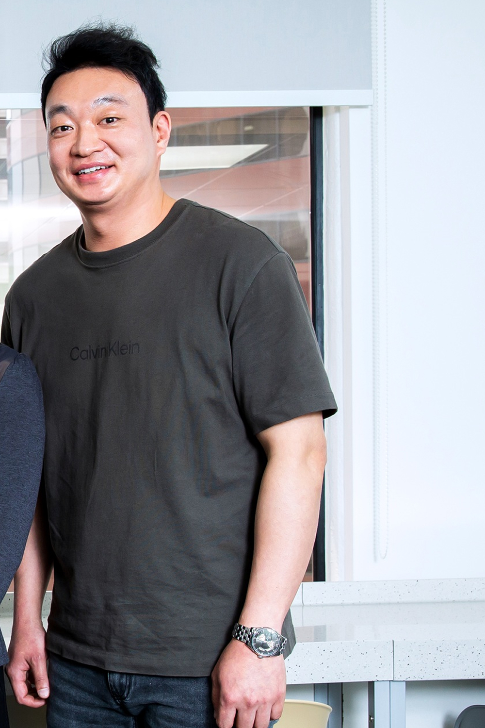 Tai Seok Kim stands indoors in front of a large window, smiling at the camera in a dark T-shirt and jeans.