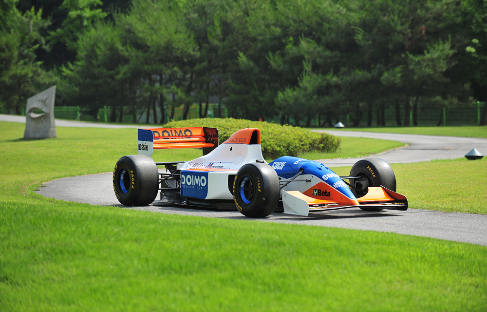 A red, white and blue Minardi M193 Formula One car is displayed on a landscaped roadway surrounded by trees.