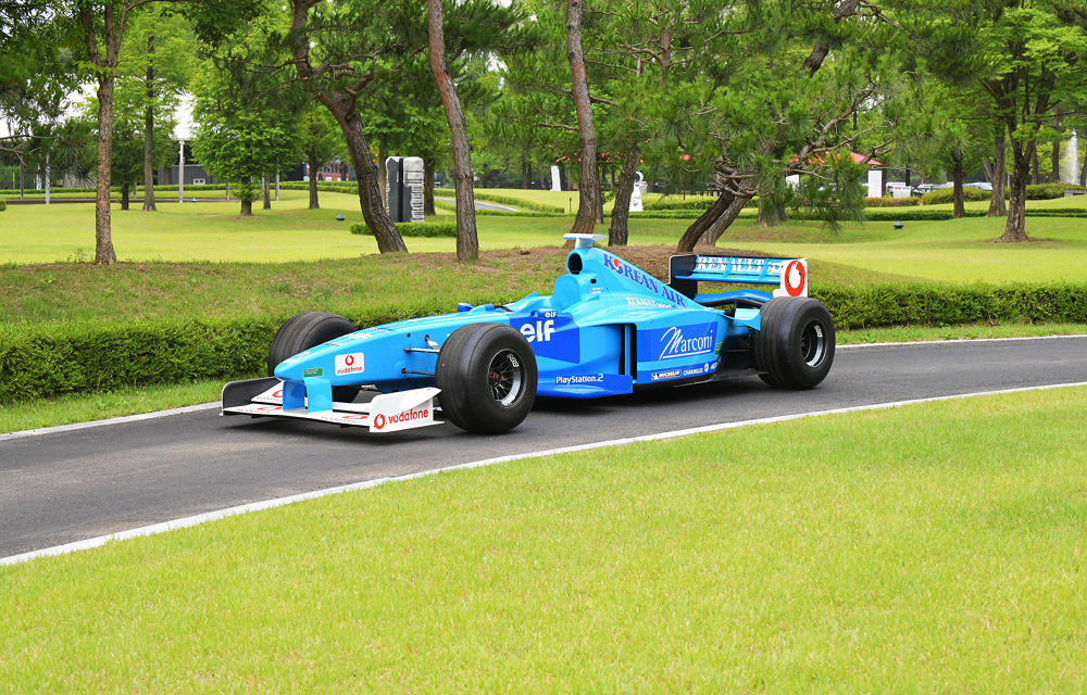 A blue Benetton B199 Formula One car is displayed outdoors on a paved road beside green grass and trees.