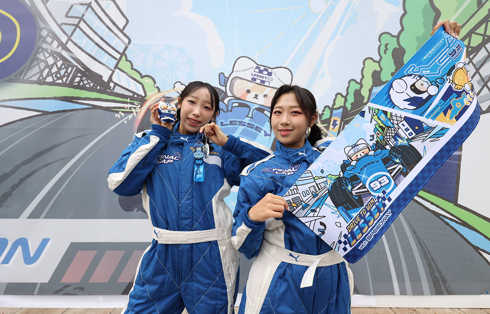 Two women wearing blue racing suits pose with a colorful race-car-shaped board in front of an illustrated racetrack backdrop.