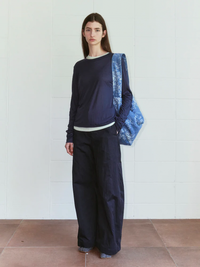 A model stands against a white tiled wall wearing a sheer navy long-sleeve T-shirt layered over a white top with dark wide-leg pants. A large blue floral-patterned bag is carried over one shoulder.