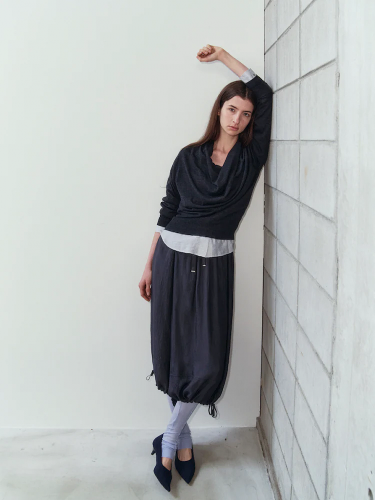 A model leans against a white tiled wall wearing a draped black cowl-neck pullover over a light gray top, paired with voluminous black satin skirt pants, pale lavender tights and black pointed heels.