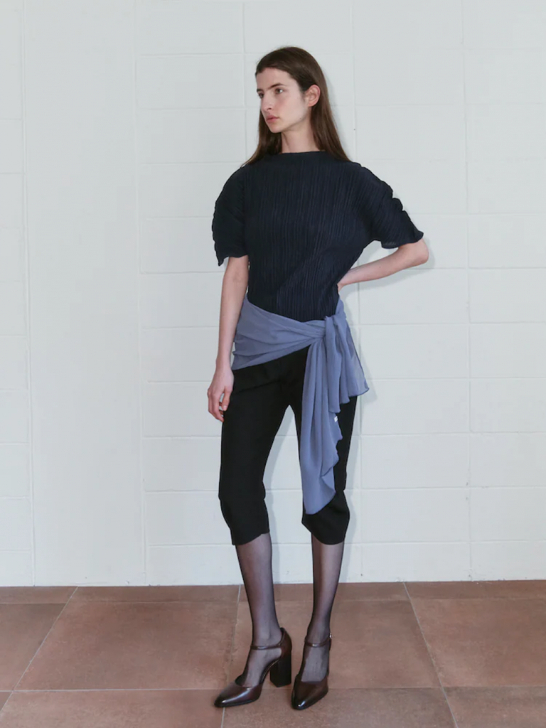 A model stands against a white tiled wall wearing a navy pleated short-sleeve top with fitted black capri pants. A light blue garment is wrapped around the waist, completing the look with sheer black socks and dark brown heels.