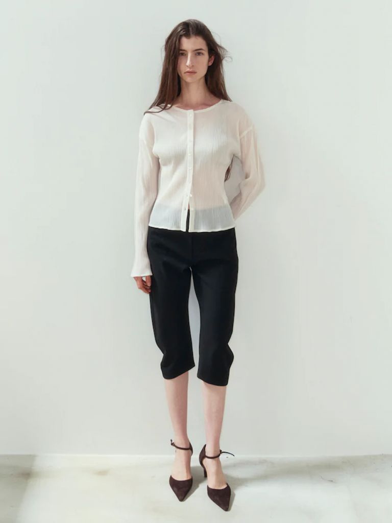 A model stands against a pale background wearing a semi-sheer ivory pleated cardigan with black capri pants and dark brown pointed heels.